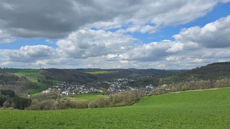 Eine grüne Hügellandschaft mit einem kleinen Dorf in der Täler. Der Himmel ist bewölkt und zeigt einige sonnige Stellen.