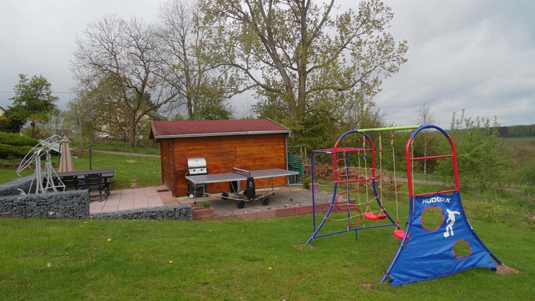 Ein Spielplatz mit verschiedenen Spielgeräten, darunter eine Schaukel und eine Rutsche. Im Hintergrund befindet sich ein Holzhaus und einige Bäume.
