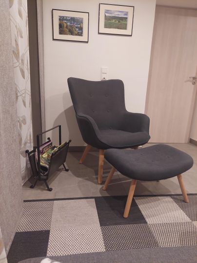 A comfortable gray armchair with a matching footstool is located in a modern room. Pictures hang on the wall, and there is a magazine holder next to the armchair.