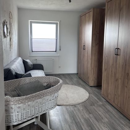 Ein heller Raum mit einem Sofa, einem Teppich und einem Babybett. Der Raum hat Holzschranktüren und ein Fenster, das natürliches Licht hereinlässt.