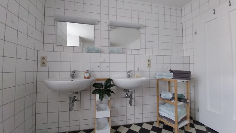 Ein modernes Badezimmer mit zwei Waschbecken und großen Spiegeln. Der Boden ist mit schwarz-weißen Fliesen gestaltet und es gibt einen Stauraum für Handtücher.