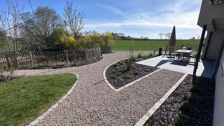 A well-maintained garden path leads through a green landscape. In the background, there is a seating area with a table and sun umbrella.