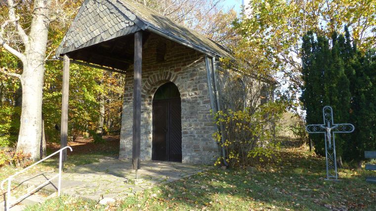 Ein kleiner Steingebäude mit einem Dach aus Schiefer steht in einem Waldgebiet. Umgeben von Bäumen und Grünflächen strahlt es eine ruhige, beschauliche Atmosphäre aus.