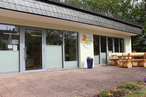 Ein modernes Gebäude mit großen Fenstern und einem einladenden Eingangsbereich. Vor dem Gebäude steht eine Holzbank in einem ruhigen Garten.