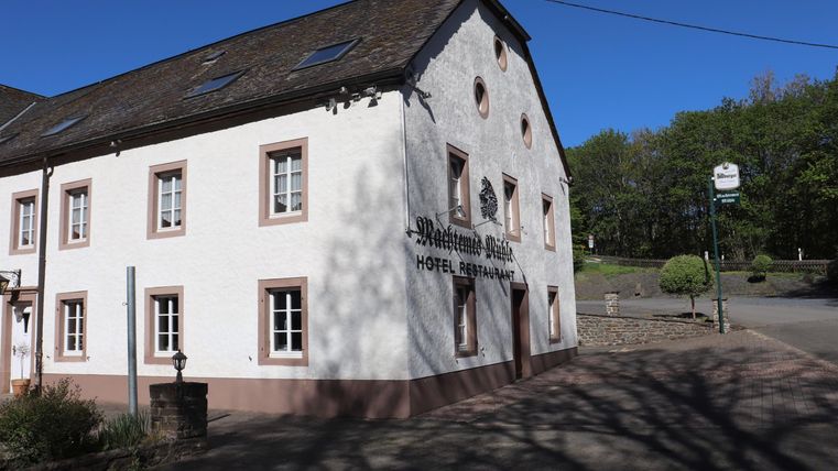 Ein historisches Gebäude mit weißer Fassade und roten Fenstern. Im Hintergrund sind Bäume und blauer Himmel zu sehen.