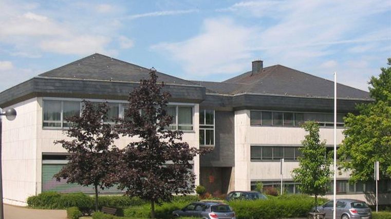 Ein modernes Gebäude mit einem Steildach und großen Fenstern. Umgeben von Bäumen und einigen geparkten Autos.