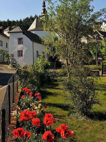Ein malerisches Dorf mit einer kleinen Kapelle und blühenden Blumen im Vordergrund. Die Umgebung ist grün und einladend, ideal für einen ruhigen Spaziergang.