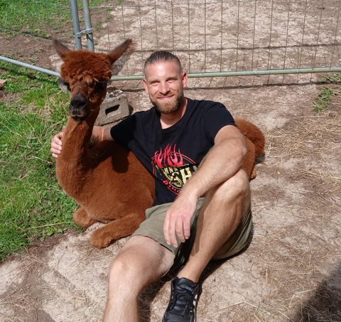 Ein Mann sitzt neben einem Alpaka auf dem Boden. Beide wirken entspannt und glücklich in einer grünen Umgebung.