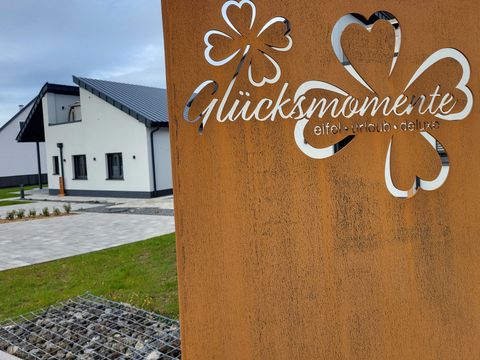 Ein moderner Gebäudekomplex mit einem rustikalen Eingangsschild, das die Worte "Glücksmomente" zeigt. Die Umgebung ist gepflegt mit grünem Gras und Steinflächen.