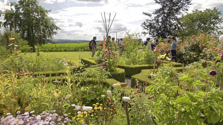 Ein schöner Garten mit bunten Blumen und einem kunstvoll gestalteten Buchsbaum-Labyrinth. Im Hintergrund sind Menschen zu sehen, die die Natur genießen.