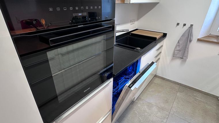 A modern kitchen with a built-in oven and a dishwasher. The surfaces are bright and shiny, the design is minimalist.