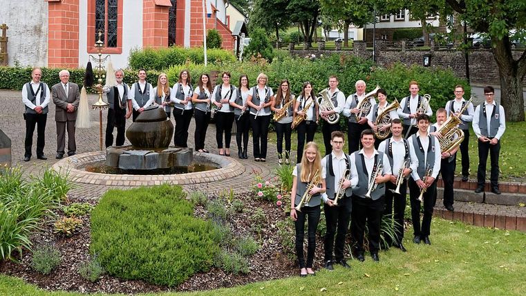 Eine Gruppe von Musikern steht im Freien vor einer Kirche und einer Wasserstelle. Sie tragen einheitliche Kleidung und halten Musikinstrumente in den Händen.