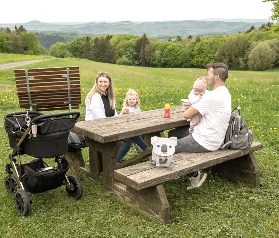 Eine Familie sitzt an einem Holztisch auf einer Wiese mit Blick auf die Hocheifel. Ein Kinderwagen steht daneben, im Hintergrund sind Wälder und Hügel zu sehen.