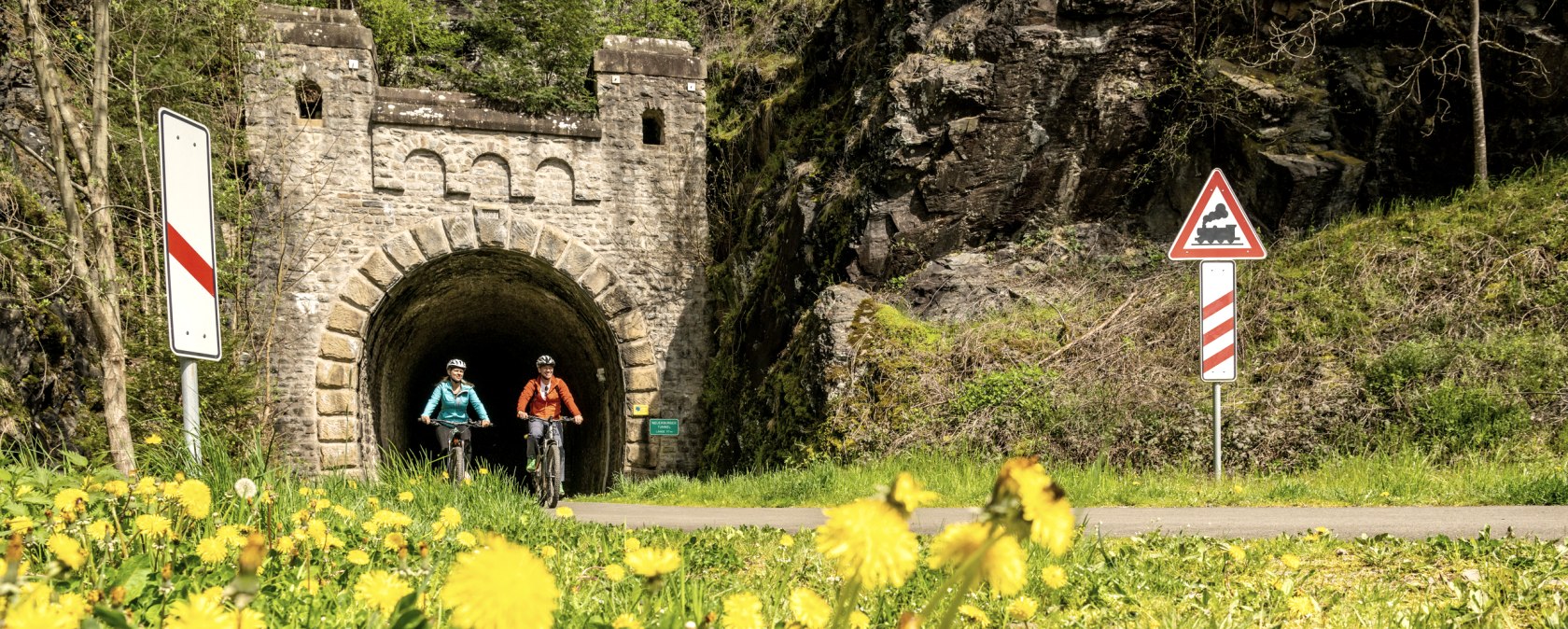 Zwei Radfahrer auf einem Weg vor einem alten Bahntunnel, umgeben von blühendem Löwenzahn und Verkehrsschildern., © Eifel Tourismus GmbH, Dominik Ketz