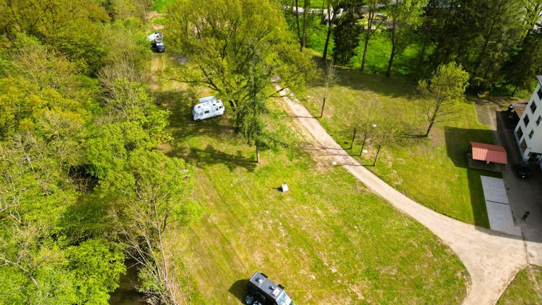 Eine grüne Wiese mit Bäumen und einem Parkplatz für Campingfahrzeuge. Im Hintergrund verläuft ein Weg und ein Gebäude ist sichtbar.