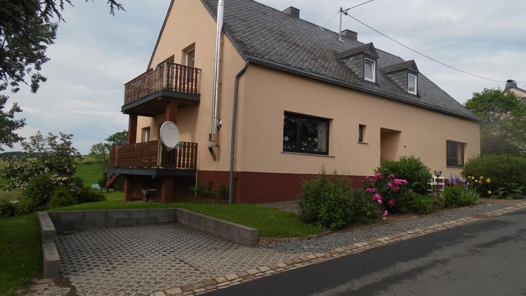 Ein schlichtes Wohnhaus mit einem Balkon und Gartenblumen. Die Auffahrt ist mit Steinen gepflastert und die Umgebung ist grün.