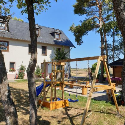 Ein Spielplatz mit einer Schaukel und einer Rutsche im Garten. Im Hintergrund steht ein weißes Haus und die Sonne scheint.