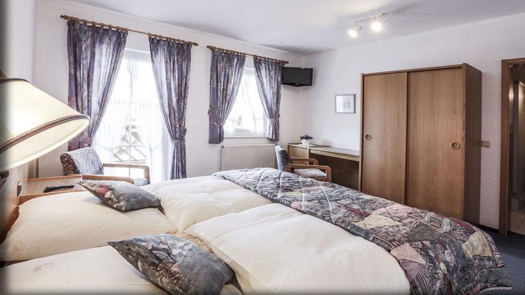 Ein gemütliches Schlafzimmer mit zwei Einzelbetten und einem Schreibtisch. Große Fenster lassen viel Tageslicht herein, und die Vorhänge sorgen für eine angenehme Atmosphäre.