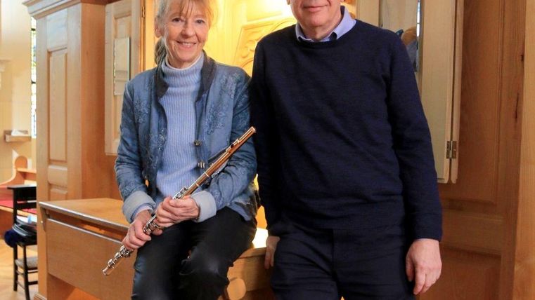 Eine Frau sitzt mit einer Flöte in der Hand, während ein Mann daneben steht. Beide lächeln und posieren vor einem musikinstrumentalen Hintergrund.