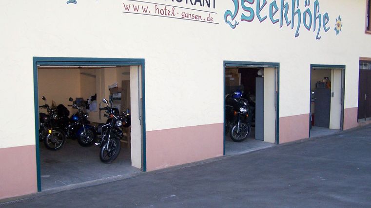 Eine Garage mit mehreren Motorrädern und einem Restaurant im Hintergrund. Die Fassade ist hell und einladend.