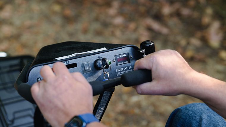 Eine Person hält einen Steuerhebel des Rollstuhl-Zuggerätes. Der Hintergrund zeigt verschwommene Natur.