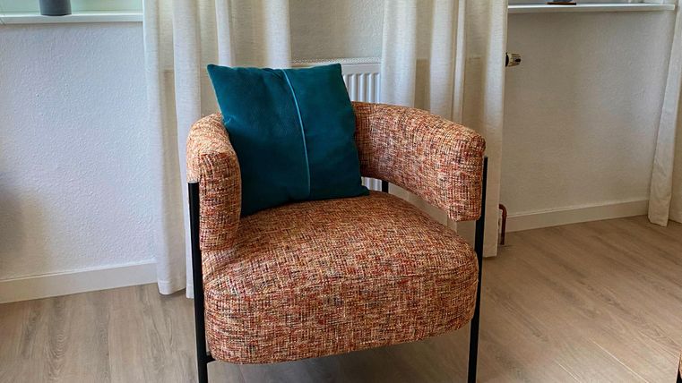 Een moderne stoel met een kleurrijke stofoppervlak en een blauwe kussen. Op de achtergrond zijn er ramen met gordijnen en een kleine plant te zien.
