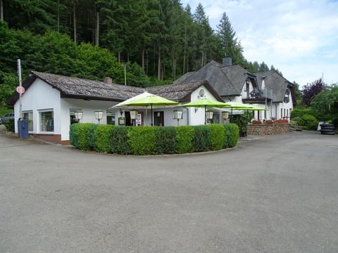 Ein idyllisches Gasthaus mit einer einladenden Terrasse und grünen Pflanzen. Im Hintergrund sind bewaldete Hügel zu sehen.