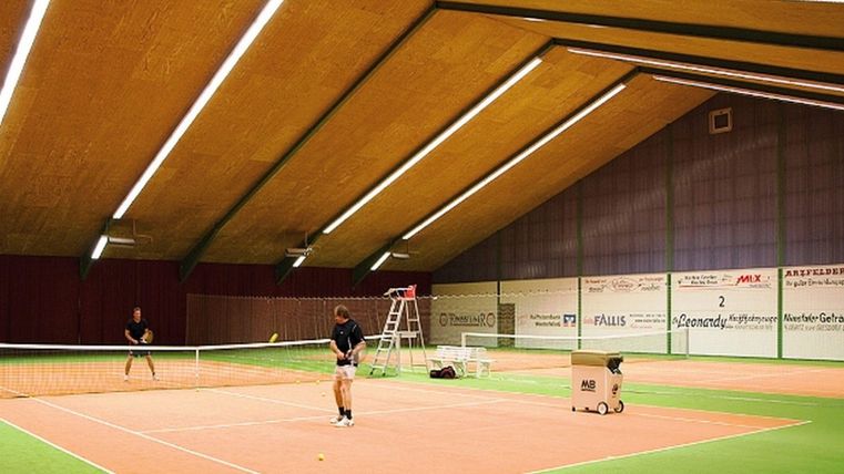 Eine indoor Tennisanlage mit zwei Spielern auf dem Platz. Das Spielfeld hat einen orangefarbenen Belag und die Wände sind mit Werbung bedeckt.