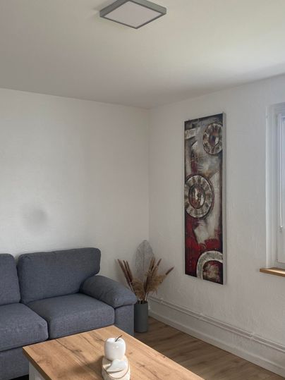 Ein einfaches Wohnzimmer mit einem grauen Sofa und einem Holztisch. An der Wand hängt ein dekoratives Bild.