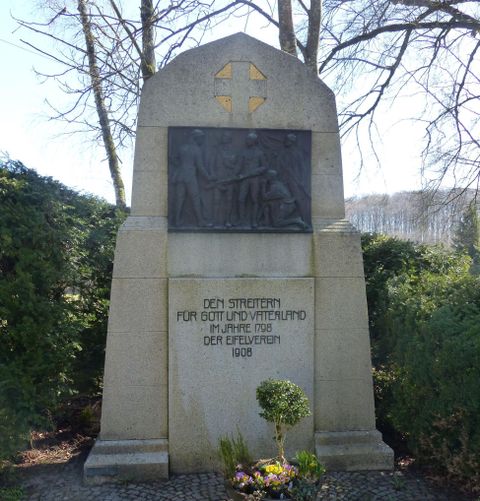 A monument made of stone with a relief figure and inscriptions. Surrounded by bushes and flowers.