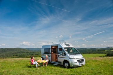 Ein Wohnmobil steht auf einer Wiese mit einer schönen Landschaft im Hintergrund. Zwei Personen sitzen entspannt draußen und genießen die Natur.