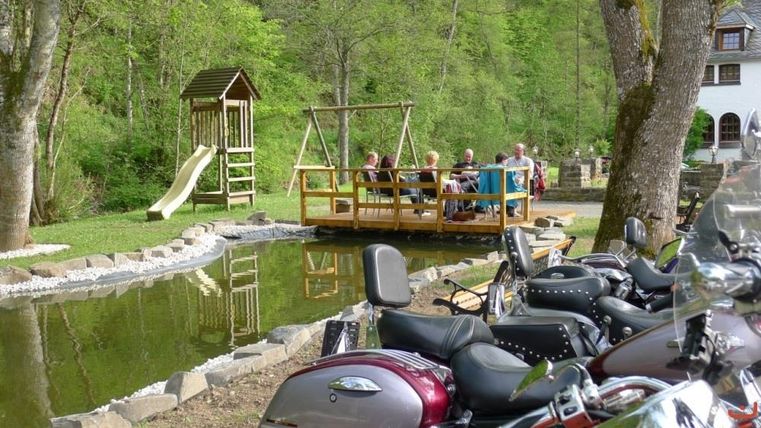 Eine entspannte Gartenlandschaft mit einem Spielplatz und einer Terrasse am Wasser. Im Vordergrund stehen Motorräder geparkt.