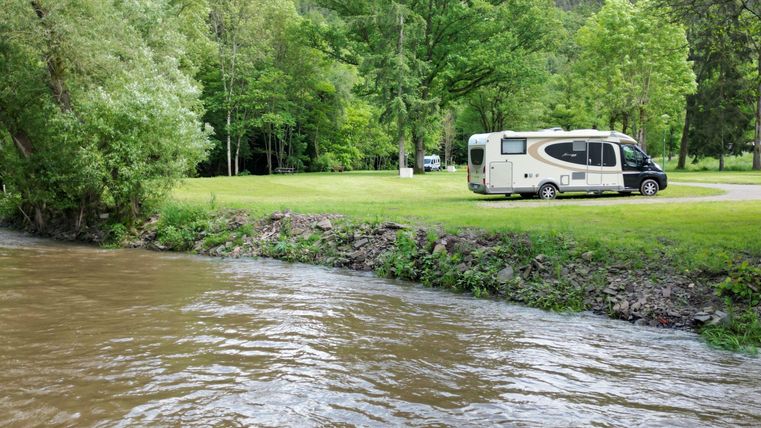 Ein Wohnmobil steht auf einer Wiese neben einem leicht strömenden Fluss. Umgeben von grünen Bäumen bietet die Szene eine ruhige, naturnahe Atmosphäre.