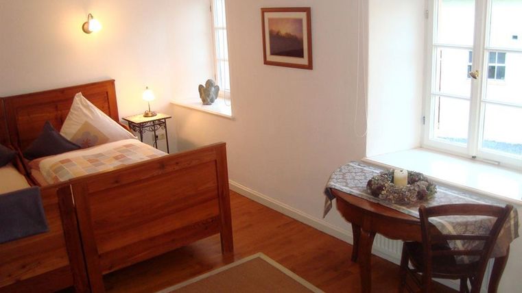 Ein gemütliches Schlafzimmer mit einem Holzbett und einem kleinen Tisch. Die Fenster lassen viel Licht hereinfliessen und schaffen eine einladende Atmosphäre.