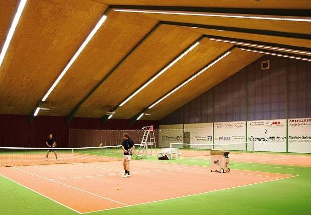 Een sporthal met een tennisbaan, waar twee spelers aan het trainen zijn. De vloer is groen en het plafond is hoog en verlicht.