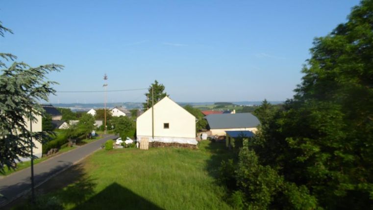 Eine ruhige Landschaft mit einem grünen Wiesenbereich und Häusern im Hintergrund. Der Himmel ist klar und blau.