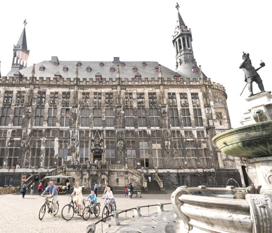 Das Aachener Rathaus mit seiner gotischen Fassade, einem Brunnen im Vordergrund und Radfahrern auf dem Platz., © vennbahn.eu