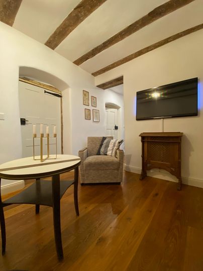 A cozy living room with an armchair, a small table, and a television on the wall. The wooden floors and ceiling beams give the room a warm atmosphere.