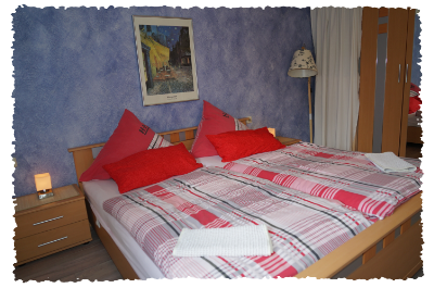 Ein gemütliches Schlafzimmer mit einem Doppelbett, roten Kissen und einer bunten Bettdecke. An der Wand hängt ein Bild, und es gibt eine Nachttischlampe sowie einen Kleiderschrank.