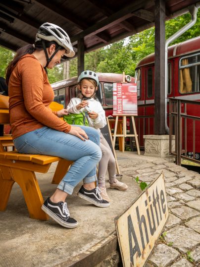 Twee mensen met fietshelmen zitten bij de Ahrhütte tijdens de Tour de Ahrtal naast een rode locomotief en een bord met de tekst „Passing Trains“.