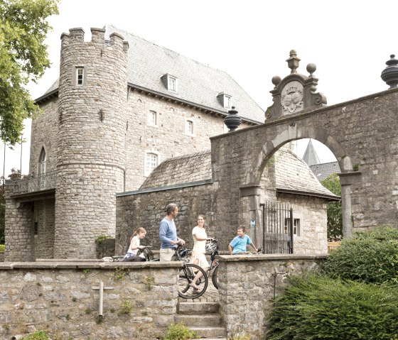 Eine Familie mit Fahrrädern steht vor dem historischen Töpfermuseum Raeren, umgeben von alten Steinmauern und einem Torbogen., © vennbahn.eu