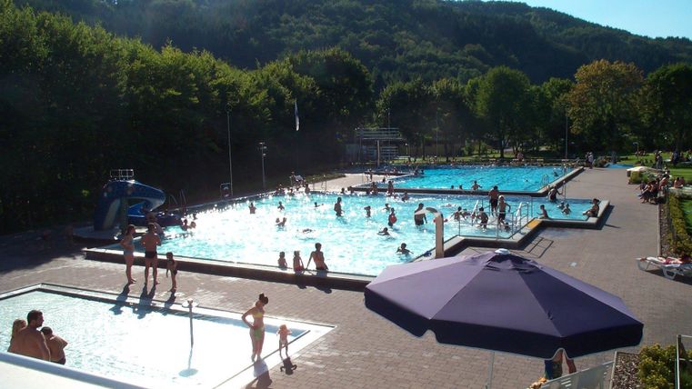 Ein Schwimmbad mit mehreren Becken und vielen Menschen, die dort schwimmen und spielen. Im Hintergrund sind grüne Hügel und Bäume zu sehen.