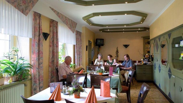 Een gezellig restaurant met vier gasten die aan een tafel zitten. De inrichting is vriendelijk, met planten en gekleurde tafelkleden.