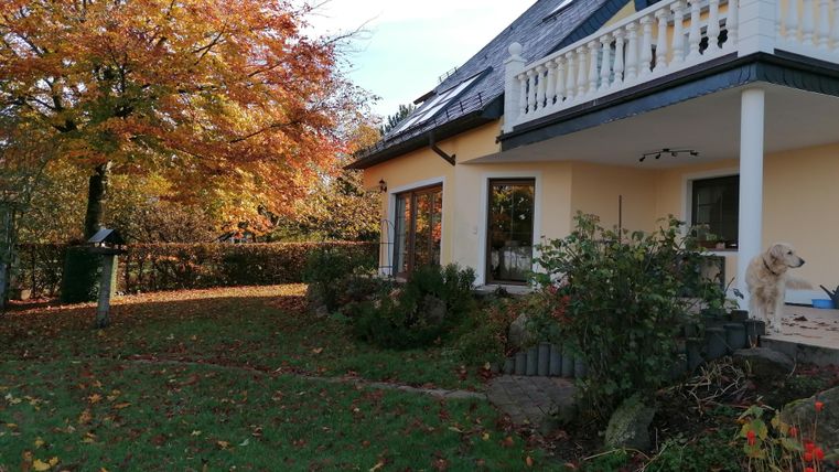 A cozy house with a balcony, surrounded by autumn trees. The garden is green and there is a dog on the terrace.