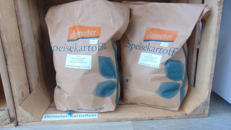 Two bags of Demeter table potatoes in a wooden box. The packaging is brown and has labels in the German language.