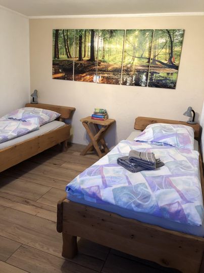 Une chambre confortable avec deux lits en bois.
Sur le mur, il y a une belle image d'une forêt, et sur la table, des serviettes sont posées.