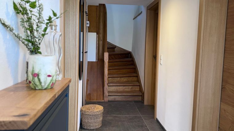 A bright hallway with a wooden staircase and modern walls. A flower vase is on a table and a basket is on the floor.