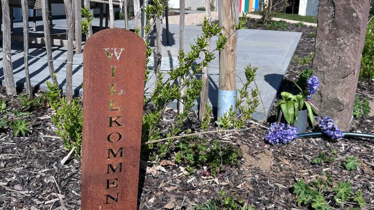 A welcome sign made of metal stands in a garden. Next to it, some plants are blooming.