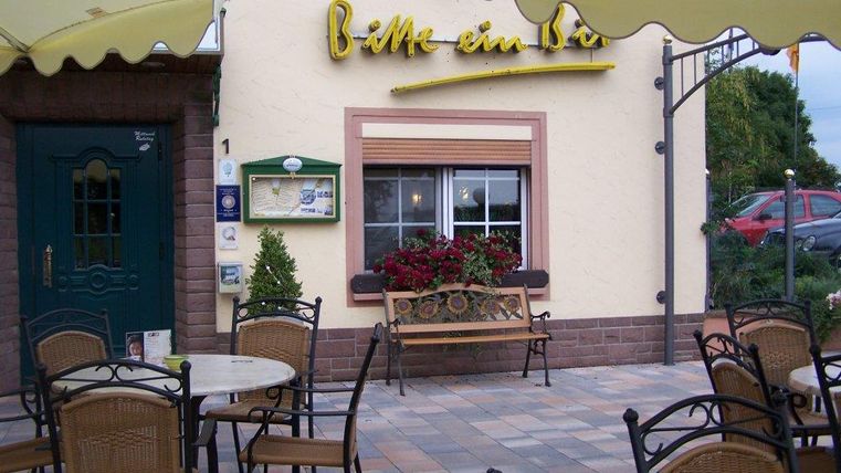 Ein einladendes Café mit Tischen und Stühlen im Freien. Die Fassade ist freundlich gestaltet und mit Blumen verziert.