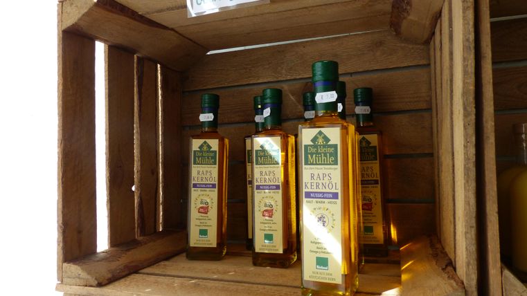 A wooden box with bottles of rapeseed oil is visible. The labels indicate organic quality and regional origin.
